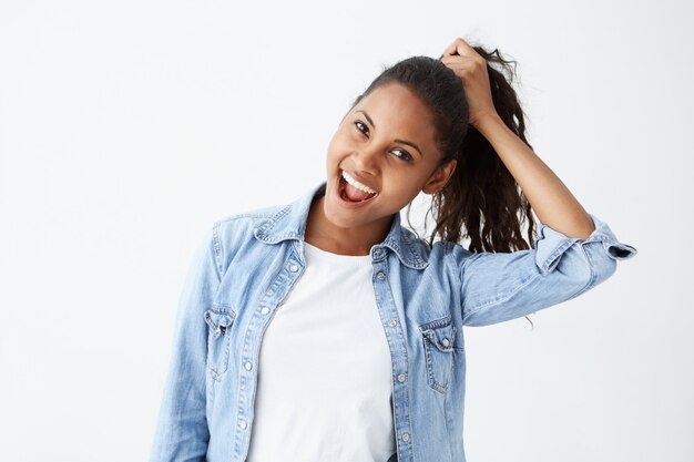 Expressões e emoções do rosto humano. Retrato de uma jovem mulher morena afro-americana em camisa jeans azul claro se divertindo, segurando o cabelo com a mão, com a boca aberta.