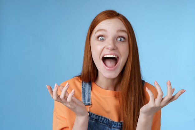 Extremamente feliz, bonita, ruiva, caucasiana, reagindo a uma notícia fantástica e incrível, gritando emocionada, levantando as mãos divertida, tremendo alegremente gritando sim, parece surpresa surpresa, fundo azul.