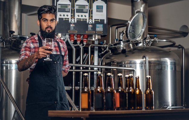 Fabricante masculino hipster tatuado e barbudo indiano degustando e apresentando cerveja artesanal na microcervejaria.