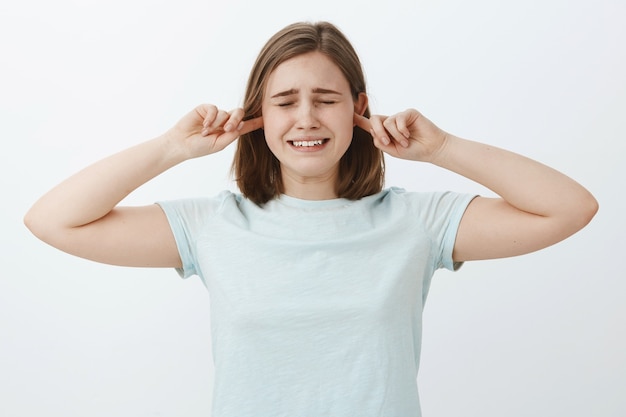 Faça esse som parar. Menina bonita infeliz e insegura chorando com cabelos castanhos fechando os olhos e cerrando os dentes, fazendo uma expressão triste e descontente cobrindo as orelhas com o dedo indicador, sem ouvir os pais brigando