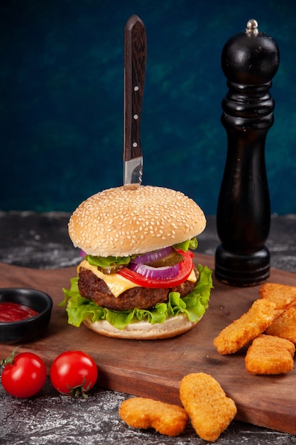 Faca no saboroso sanduíche de carne e tomate nuggets de frango com pimenta do caule na tábua de madeira molho de ketchup na superfície azul escuro