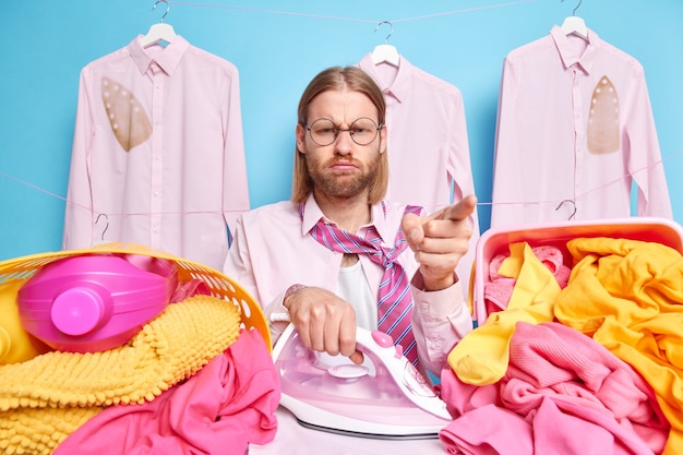 Fadiga dono da casa ocupado passando roupa em casa posa com cestos de ferro elétricos cheios de roupa suja e detergentes, usa gravata de camisa, expressão carrancuda isolada em fundo azul