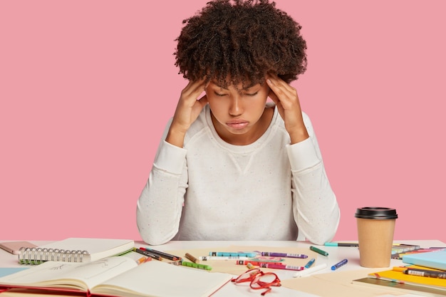 Fadiga Mulher afro-americana mantém as mãos nas têmporas, sofre de enxaqueca, suspira de cansaço, trabalha muito, posa na mesa com bloco de notas em espiral, café para viagem, isolado na parede rosa