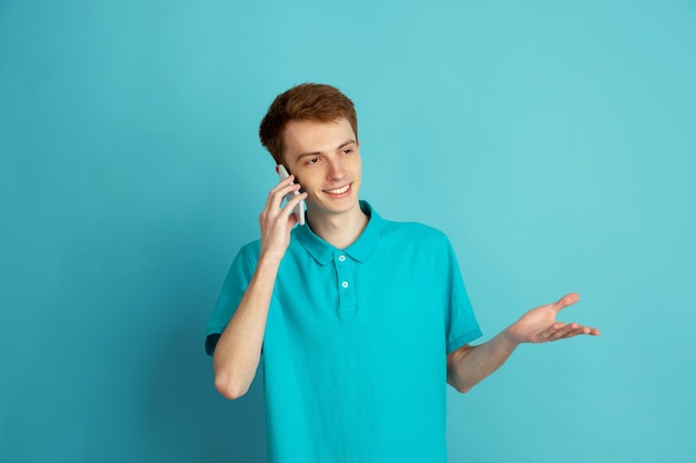 Falando ao telefone, sorrindo. Retrato moderno de jovem caucasiano no estúdio azul, monocromático.