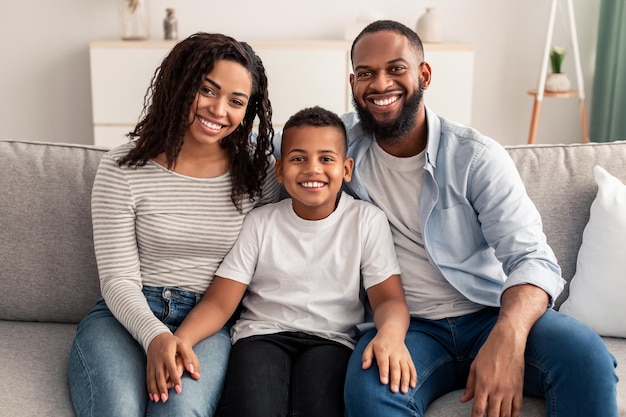 Família amorosa feliz. retrato de alegre filho afro-americano sentado no sofá entre seus pais sorridentes. mulher positiva, homem e menino posando para foto e olhando para a câmera em casa