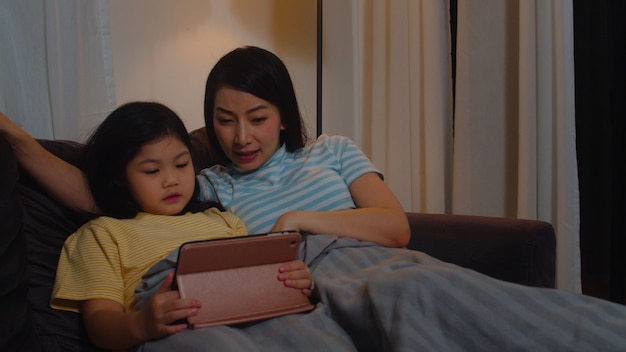 Família asiática nova e filha felizes usando a tabuleta em casa. mãe coreana relaxar com a menina assistindo filme deitado no sofá antes de ir dormir na sala de estar em casa moderna à noite.