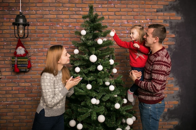 Família com pequena filha pendurada brinquedos em uma árvore de Natal