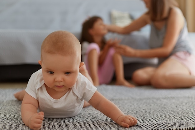 Família, criança e paternidade. Retrato de uma jovem mãe sentada com a filha mais velha sentada perto do sofá, bebê rastejando no chão, família posando em casa.