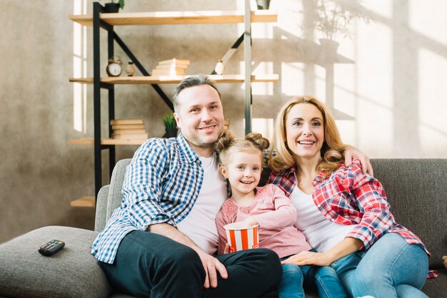 Família feliz assistindo televisão com filha comendo pipocas no sofá em casa