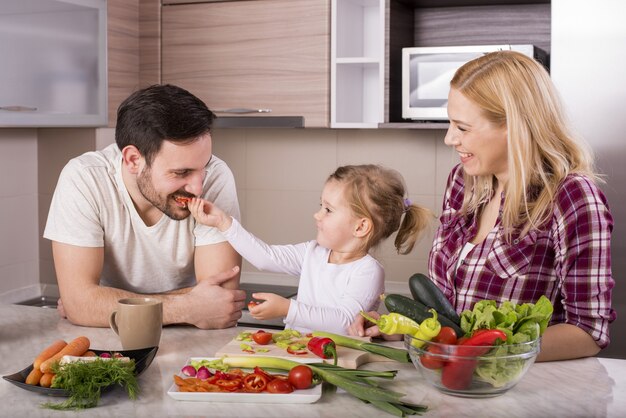 Família feliz com sua filha fazendo uma salada fresca com legumes na cozinha