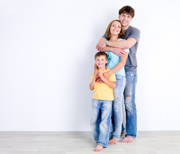 Família feliz de três pessoas se abraçando perto da parede vazia