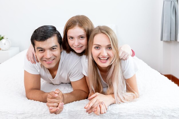 Família feliz, incluindo pai mãe e filha estão deitados na cama com sorriso olhando para a câmera