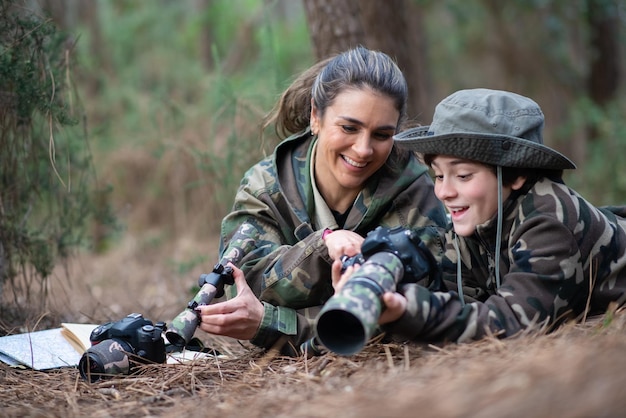 Família interessada tirando fotos na floresta. Mãe e filho com câmeras modernas deitado no chão, segurando câmeras, olhando fotos. Parentalidade, família, conceito de lazer