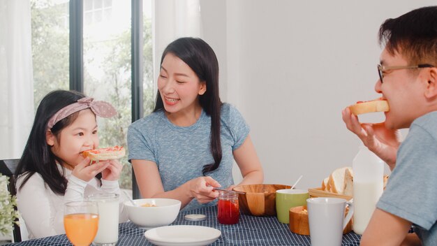 Família japonesa asiática toma café da manhã em casa. A mãe feliz asiática que faz o doce de morango no pão para a filha come flocos de milho cereal e leite na bacia na tabela na cozinha da manhã.
