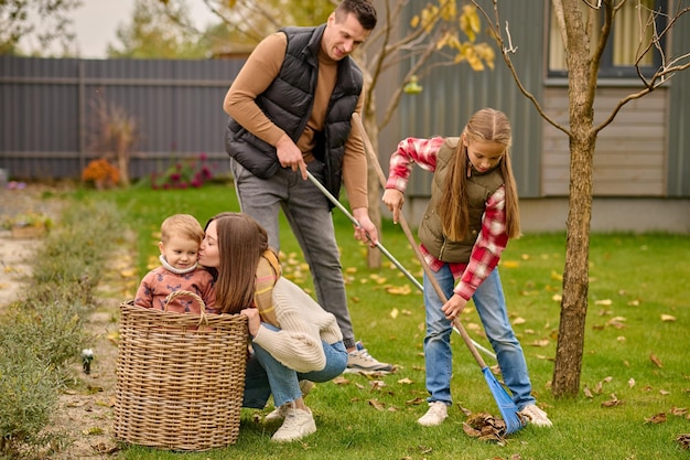 Família, jardim. homem sorridente com menina em idade escolar raking deixa uma jovem loira agachada beijando uma criança fofa na cesta no jardim no dia do outono