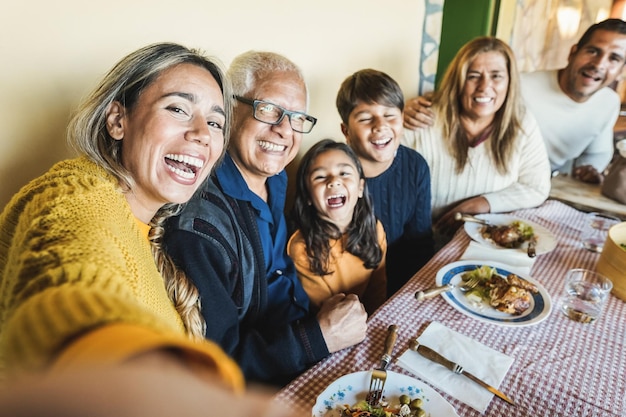 Família latina feliz fazendo selfie enquanto comem juntos em casa - foco no rosto da mãe