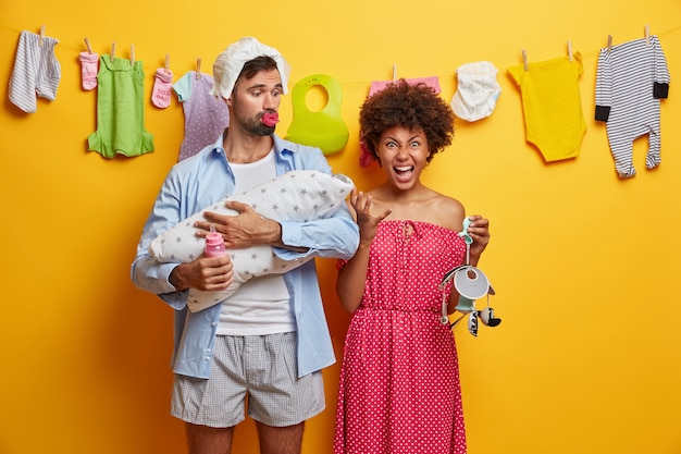 Família multirracial amigável se preocupa com o recém-nascido. pai, mãe e bebê posam em casa, alimentam e brincam com o bebê, mãe emocional irritada segura o celular pai afetuoso acalma a criança