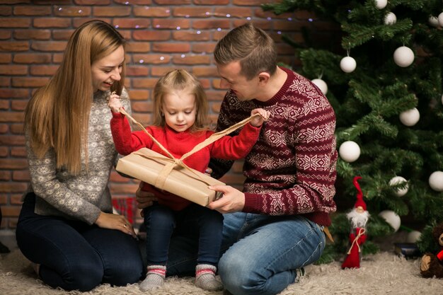 Família sentada junto à árvore de Natal com presentes de embalagem da filha pequena