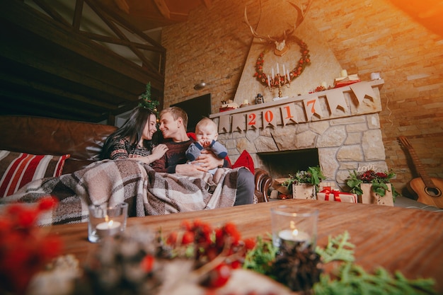 Família sentada no sofá revestido com um cobertor e visto a partir das decorações de natal da mesa de madeira
