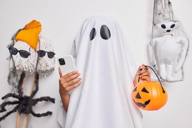 Fantasma irreconhecível segurando um celular moderno e preparações de abóbora esculpidas para pesquisas de comemoração do Dia das Bruxas em ideias de internet para decorar o quarto antes de poses de festa perto de brinquedos assustadores dentro de casa.