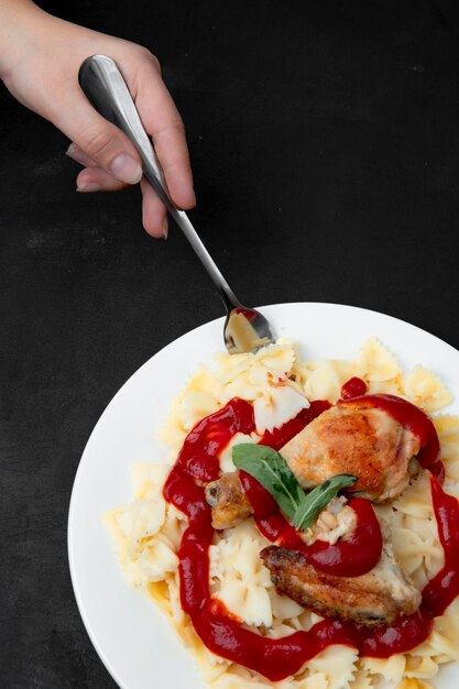 Farfalle com perna de frango e mão de uma mulher segurando o garfo na superfície preta