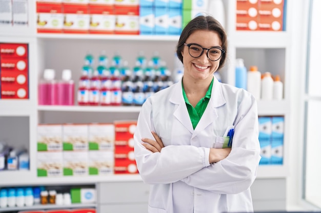 Farmacêutica jovem hispânica sorrindo confiante em pé com gesto de braços cruzados na farmácia