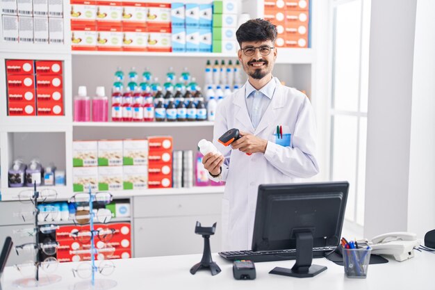 Farmacêutico jovem hispânico sorrindo confiante escaneando o frasco de comprimidos na farmácia