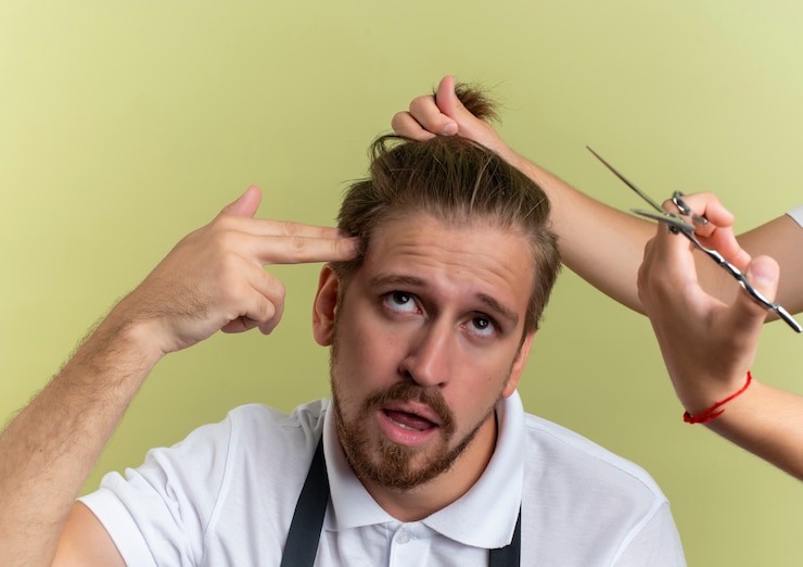 Foto grátis farto de jovem bonito barbeiro gesticulando suicídio olhando para cima com alguém segurando e se preparando para cortar o cabelo isolado em verde oliva