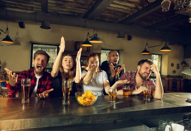 Fãs de esporte torcendo em bar, pub. Tinir copos de cerveja enquanto assiste ao campeonato, competição. Grupo multiétnico de amigos entusiasmados com a tradução. Emoções humanas, expressão, conceito de apoio.