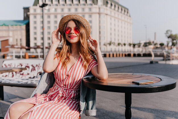 Fascinante garota branca com chapéu de palha, sentado em uma cafeteria ao ar livre. Retrato de senhora Europeia alegre em lindo vestido listrado relaxando na cidade.