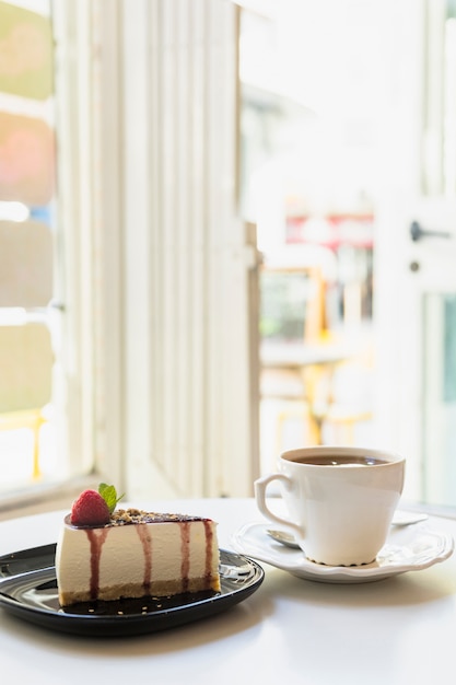 Fatia de cheesecake delicioso e xícara de chá na mesa branca perto de uma porta aberta