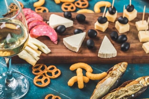 Fatias de calabresa, queijo e azeitonas pretas em uma placa de madeira com biscoitos e um copo de vinho branco