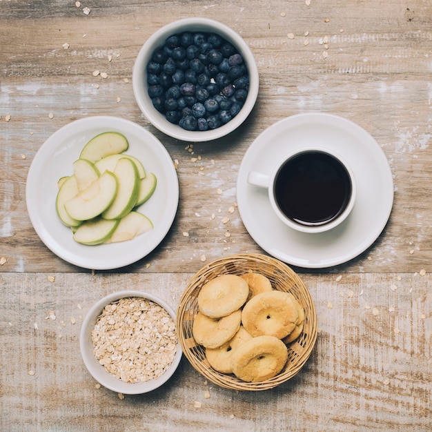 Fatias de maçã; tigela de aveia; biscoitos; tigela de mirtilo e xícara de café na madeira texturizada