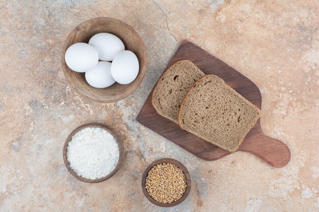 Fatias de pão, tigelas de ovos, farinha e cevada em superfície de mármore