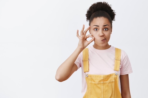 Foto grátis fechando minha boca para sempre, segredo seguro. retrato de uma mulher afro-americana séria e charmosa, fazendo uma promessa, fechando os lábios com um zíper invisível, fazendo beicinho na boca e segurando o dedo perto da bochecha