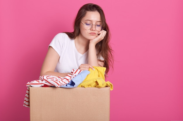 Fechar o retrato de mulher jovem infeliz posando com caixa de doação de roupas, tem expressão facial triste