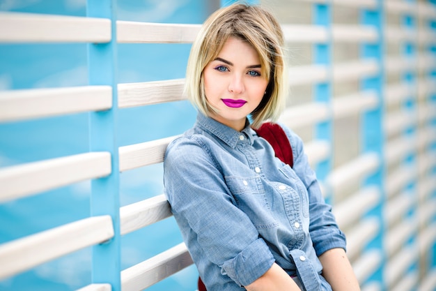 Fechar o retrato de uma menina sorridente com cabelo loiro curto, lábios rosados brilhantes e maquiagem nude encostada em uma cerca de listras azuis e brancas no fundo