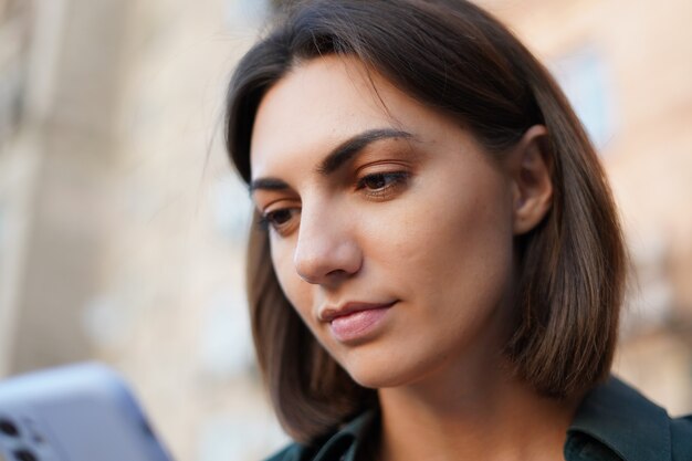 Fechar o retrato de uma mulher na rua ao pôr do sol segurando o telefone celular, olhando para a tela com um sorriso