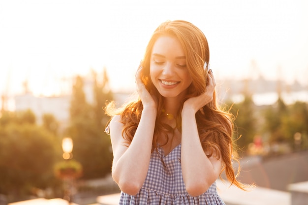 Feche a foto da mulher atraente gengibre no vestido ouvindo música por do sol e olhando para baixo