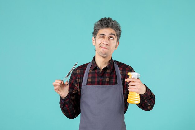 Feche a foto de um barbeiro confuso usando avental cinza e segurando um frasco de spray de tesoura na superfície de cor azul suave