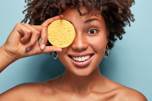 Feche a foto de uma mulher alegre com pele escura e saudável, segura a esponja no olho, tem procedimentos cosméticos, posa seminua sobre a parede azul. Mulher sorridente tem beleza natural, remove maquiagem.