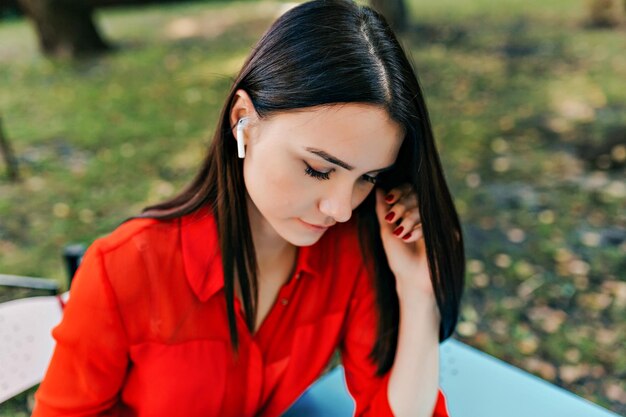 Feche a foto externa de uma mulher de cabelos escuros usando blusa vermelha e ouvindo música no terraço ao ar livre