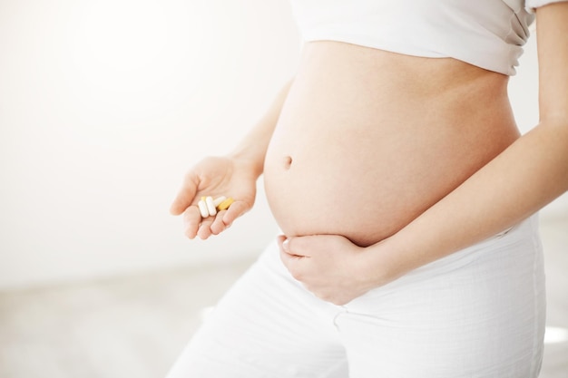 Feche a mãe em breve feliz segurando a barriga e pílulas pré-natais e vitaminas e minerais esperando por um bebê saudável Conceito pré-natal Chave alta