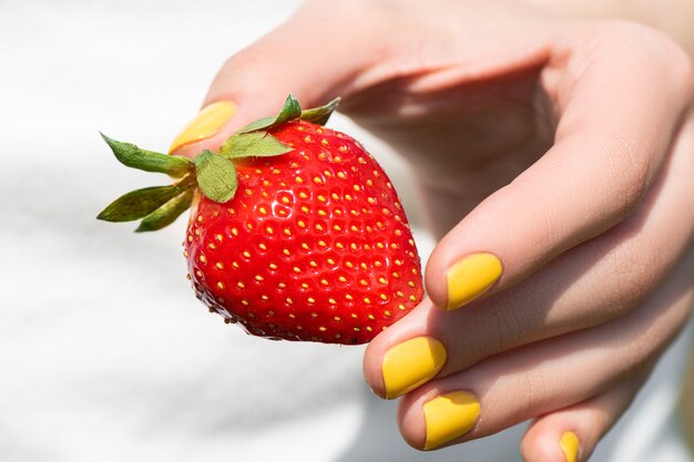 Feche a mão feminina com manicure de design de unhas muito amarelo segurando morango maduro.