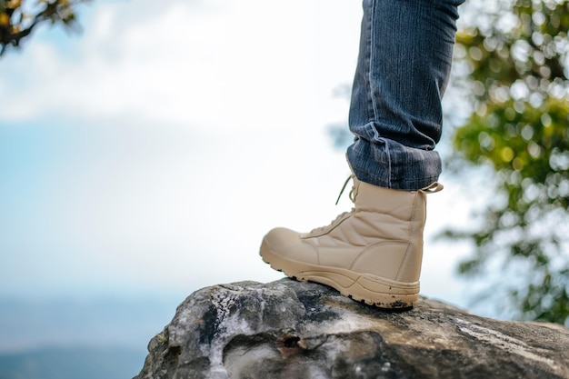 Feche a perna do homem de caminhada em jeans com tênis de trekking colocado no pico da montanha rochosa no céu azul da floresta natural no espaço de cópia de fundo