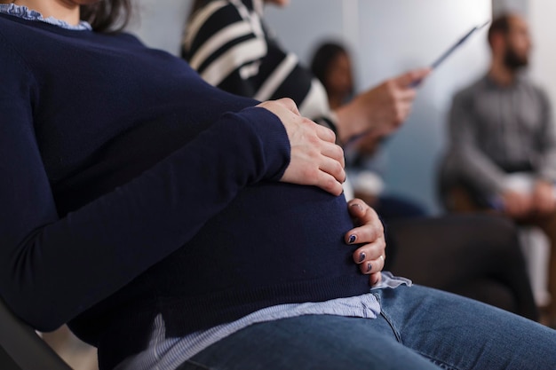 Foto grátis feche a recrutadora de emprego grávida segurando a mão na barriga esperando na área do saguão do escritório de negócios de inicialização. grupo multiétnico de candidatos lendo currículo cv antes da reunião da entrevista