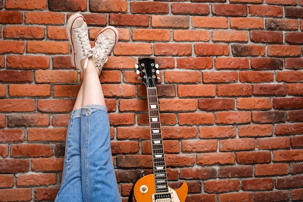 Feche acima das pernas e da guitarra da menina sobre o fundo do tijolo.