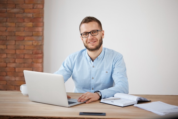 Feche acima do gerente masculino barbudo adulto alegre nos vidros e na camisa clássica que sentam-se na mesa no escritório, trabalhando no computador pessoal, escrevendo a informação no caderno.