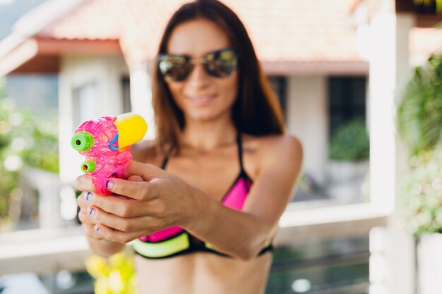 Feche as mãos de uma mulher muito sorridente e feliz brincando com um revólver de água na piscina nas férias de verão tropical em um hotel villa se divertindo em biquínis, estilo colorido, clima de festa