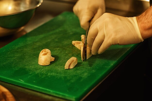Feche as mãos do chef em luvas brancas segurando o chef de faca cortando cogumelos no conceito de tábua verde de cozinha de cozinha de restaurante e culinária